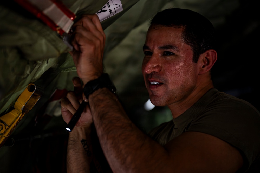 A man in a military-style olive green shirt is working in a confined, dimly lit space, appearing to inspect or maintain mechanical components. He is holding a small flashlight and manipulating what appears to be safety-tagged equipment with red and white warning markings. His face shows concentration and perspiration, suggesting physical exertion in a warm environment.