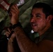 A man in a military-style olive green shirt is working in a confined, dimly lit space, appearing to inspect or maintain mechanical components. He is holding a small flashlight and manipulating what appears to be safety-tagged equipment with red and white warning markings. His face shows concentration and perspiration, suggesting physical exertion in a warm environment.