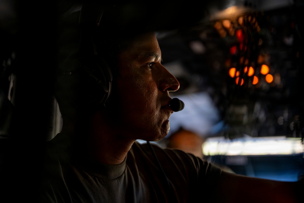 A person wearing aviation headset with boom microphone is photographed in dramatic side-profile lighting inside what appears to be a military or commercial aircraft cockpit. The image uses strong chiaroscuro lighting, with the subject's face partially illuminated against a dark background. Illuminated instrument panels and cockpit controls are visible in the background.