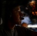 A person wearing aviation headset with boom microphone is photographed in dramatic side-profile lighting inside what appears to be a military or commercial aircraft cockpit. The image uses strong chiaroscuro lighting, with the subject's face partially illuminated against a dark background. Illuminated instrument panels and cockpit controls are visible in the background.