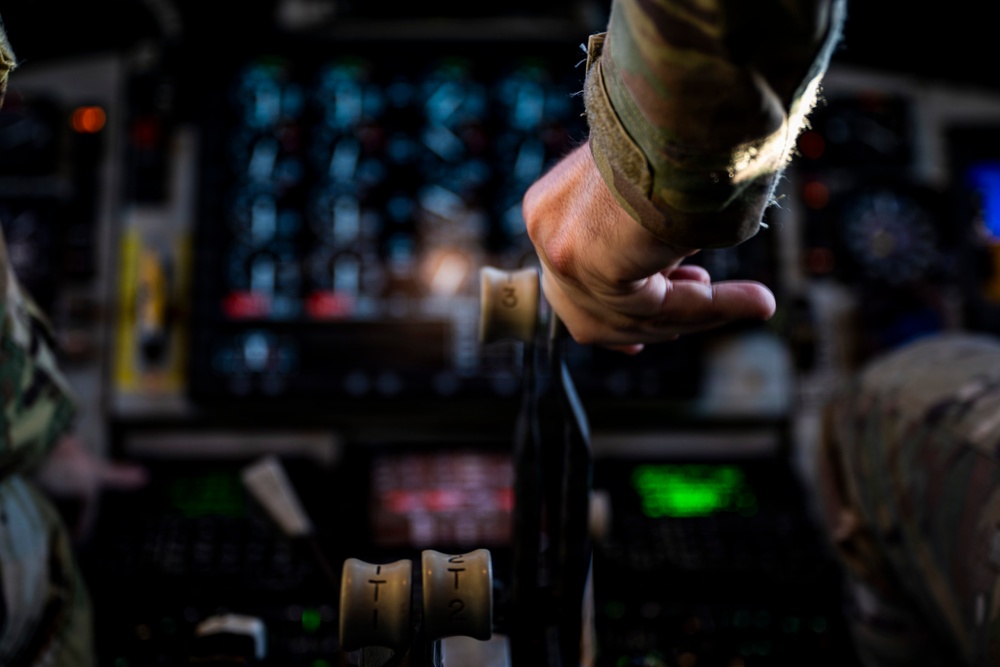 A military aircrew member in camouflage uniform grips what appears to be a throttle or control lever inside a military aircraft cockpit. The cockpit is filled with numerous illuminated instruments, switches, and displays visible in the background. The image is taken in low-light conditions with a shallow depth of field, keeping the hand and control in sharp focus.