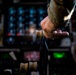 A military aircrew member in camouflage uniform grips what appears to be a throttle or control lever inside a military aircraft cockpit. The cockpit is filled with numerous illuminated instruments, switches, and displays visible in the background. The image is taken in low-light conditions with a shallow depth of field, keeping the hand and control in sharp focus.