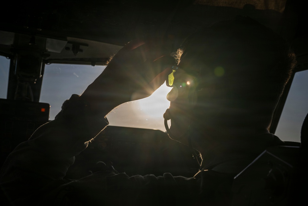 A silhouetted individual is seated inside what appears to be a vehicle or aircraft cockpit, backlit by intense sunlight creating strong lens flare effects. The person appears to be inserting or adjusting an earplug near their ear. The dramatic backlighting renders most details in deep shadow, creating a high-contrast artistic composition.
