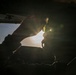 A silhouetted individual is seated inside what appears to be a vehicle or aircraft cockpit, backlit by intense sunlight creating strong lens flare effects. The person appears to be inserting or adjusting an earplug near their ear. The dramatic backlighting renders most details in deep shadow, creating a high-contrast artistic composition.