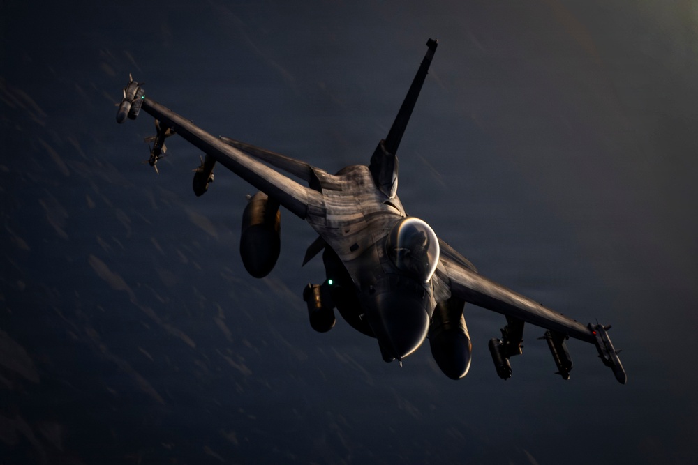 A military fighter jet, consistent with an F-16 Fighting Falcon, is photographed in flight from a slightly elevated frontal angle against a dark, cloud-streaked sky. The aircraft is heavily loaded with multiple external stores including missiles and fuel tanks. Low-angle dramatic lighting illuminates the underside and nose of the aircraft, creating strong contrast against the dark background.