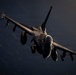 A military fighter jet, consistent with an F-16 Fighting Falcon, is photographed in flight from a slightly elevated frontal angle against a dark, cloud-streaked sky. The aircraft is heavily loaded with multiple external stores including missiles and fuel tanks. Low-angle dramatic lighting illuminates the underside and nose of the aircraft, creating strong contrast against the dark background.