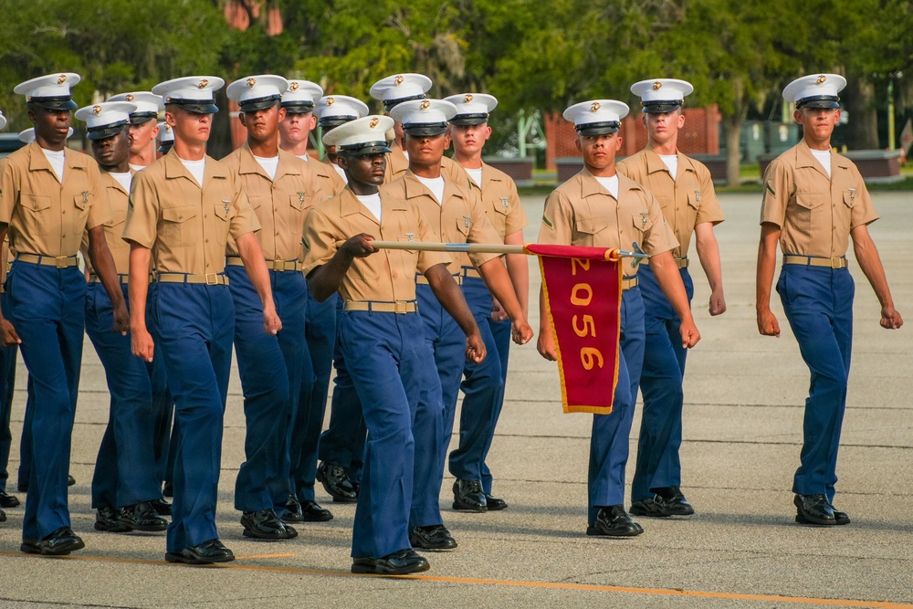 A formation of U.S. Marine Corps recruits or graduates marches in a parade drill on a paved parade deck. One individual carries a red guidon pennant bearing the number '2056', identifying their platoon or company unit. The Marines are dressed in Service Charlie uniform consisting of khaki shirts, blue trousers, and white covers (caps).