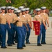 A formation of U.S. Marine Corps recruits or graduates marches in a parade drill on a paved parade deck. One individual carries a red guidon pennant bearing the number '2056', identifying their platoon or company unit. The Marines are dressed in Service Charlie uniform consisting of khaki shirts, blue trousers, and white covers (caps).