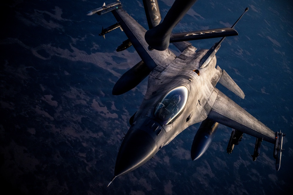 An F-16 Fighting Falcon jet fighter is captured in aerial refueling operation, receiving fuel from a tanker aircraft via a boom refueling system. The image is taken from the perspective of the tanker aircraft looking down at the fighter jet, with terrain visible far below. Dramatic low-angle lighting highlights the aircraft's metallic surfaces and weapons loadout.