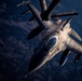 An F-16 Fighting Falcon jet fighter is captured in aerial refueling operation, receiving fuel from a tanker aircraft via a boom refueling system. The image is taken from the perspective of the tanker aircraft looking down at the fighter jet, with terrain visible far below. Dramatic low-angle lighting highlights the aircraft's metallic surfaces and weapons loadout.