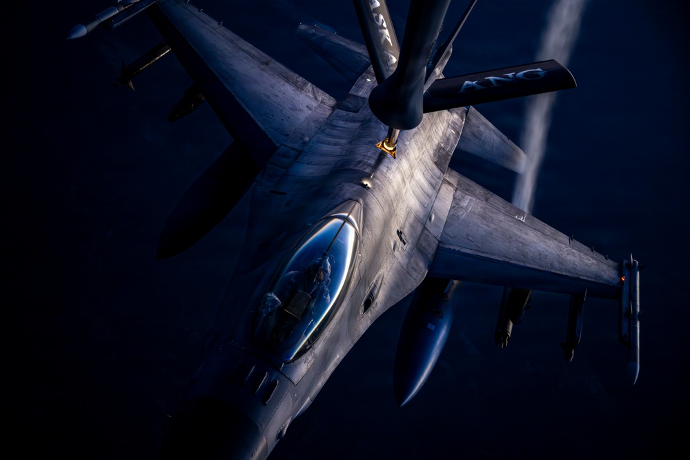 An aerial refueling operation is captured in dramatic low-light conditions, showing a fighter jet — consistent with an F-16 Fighting Falcon — receiving fuel from a tanker aircraft via a boom refueling system. The image is taken from the perspective of the tanker aircraft looking down at the receiver jet. The photograph features strong directional lighting that illuminates the aircraft's fuselage against a very dark sky background.