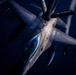 An aerial refueling operation is captured in dramatic low-light conditions, showing a fighter jet — consistent with an F-16 Fighting Falcon — receiving fuel from a tanker aircraft via a boom refueling system. The image is taken from the perspective of the tanker aircraft looking down at the receiver jet. The photograph features strong directional lighting that illuminates the aircraft's fuselage against a very dark sky background.