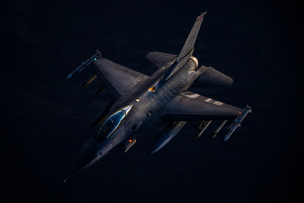 An F-16 Fighting Falcon jet fighter is photographed from above at an oblique angle during flight, likely at dusk or dawn given the dramatic low-angle lighting. The aircraft is armed with multiple air-to-air and air-to-ground munitions visible on its wing hardpoints. The tail markings suggest affiliation with the 52nd Fighter Wing of the United States Air Force.