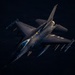 An F-16 Fighting Falcon jet fighter is photographed from above at an oblique angle during flight, likely at dusk or dawn given the dramatic low-angle lighting. The aircraft is armed with multiple air-to-air and air-to-ground munitions visible on its wing hardpoints. The tail markings suggest affiliation with the 52nd Fighter Wing of the United States Air Force.