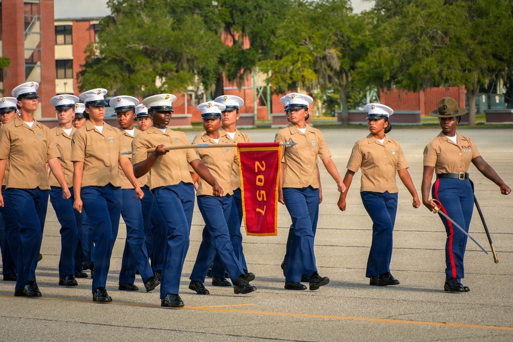 A formation of female U.S. Marine Corps recruits or graduates marches in a military parade or graduation ceremony. They are wearing Service Charlie uniforms with khaki shirts and blue trousers, led by a Drill Instructor in a campaign cover (drill sergeant hat) carrying a saber. A guidon bearer carries a red pennant marked '2057', identifying their platoon.