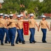 A formation of female U.S. Marine Corps recruits or graduates marches in a military parade or graduation ceremony. They are wearing Service Charlie uniforms with khaki shirts and blue trousers, led by a Drill Instructor in a campaign cover (drill sergeant hat) carrying a saber. A guidon bearer carries a red pennant marked '2057', identifying their platoon.