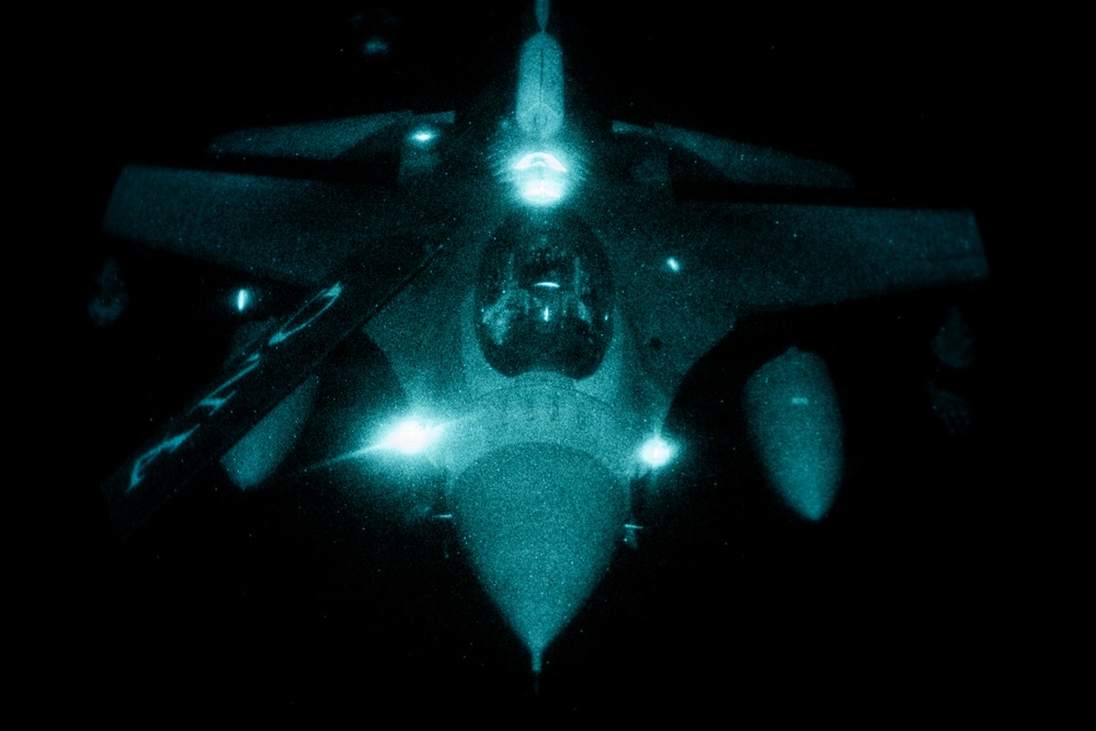 A military jet aircraft is photographed from above at night, captured through what appears to be night-vision or infrared imaging equipment, giving the image a distinctive teal/green monochromatic appearance. The aircraft is viewed from a top-down or slightly forward angle, likely taken from a refueling tanker aircraft during an aerial refueling operation. Multiple navigation and formation lights are visible and glowing brightly against the dark background.