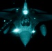 A military jet aircraft is photographed from above at night, captured through what appears to be night-vision or infrared imaging equipment, giving the image a distinctive teal/green monochromatic appearance. The aircraft is viewed from a top-down or slightly forward angle, likely taken from a refueling tanker aircraft during an aerial refueling operation. Multiple navigation and formation lights are visible and glowing brightly against the dark background.