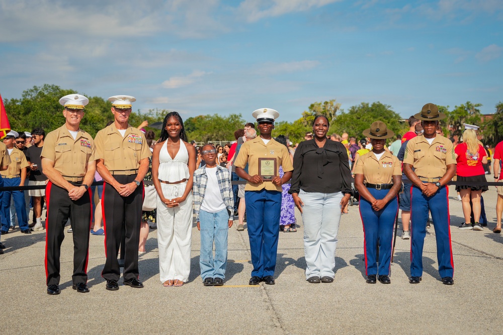 A group of seven individuals pose for a formal photograph at what appears to be a U.S. Marine Corps ceremony or graduation event. The group includes several Marines in service uniforms and two civilians, with one Marine holding a wooden award plaque. The setting is an outdoor parade deck or tarmac with a crowd of spectators visible in the background.