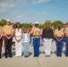 A group of seven individuals pose for a formal photograph at what appears to be a U.S. Marine Corps ceremony or graduation event. The group includes several Marines in service uniforms and two civilians, with one Marine holding a wooden award plaque. The setting is an outdoor parade deck or tarmac with a crowd of spectators visible in the background.