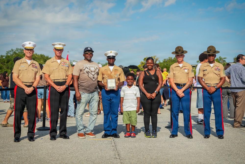 A group photo at what appears to be a U.S. Marine Corps ceremony or event, featuring uniformed Marines alongside civilians including a child. Several Marines are wearing dress uniforms with distinctive covers (white officer caps and olive drill instructor hats), and one Marine is holding a plaque or certificate. The setting appears to be an outdoor military installation or public event with a crowd visible in the background.