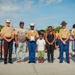 A group photo at what appears to be a U.S. Marine Corps ceremony or event, featuring uniformed Marines alongside civilians including a child. Several Marines are wearing dress uniforms with distinctive covers (white officer caps and olive drill instructor hats), and one Marine is holding a plaque or certificate. The setting appears to be an outdoor military installation or public event with a crowd visible in the background.