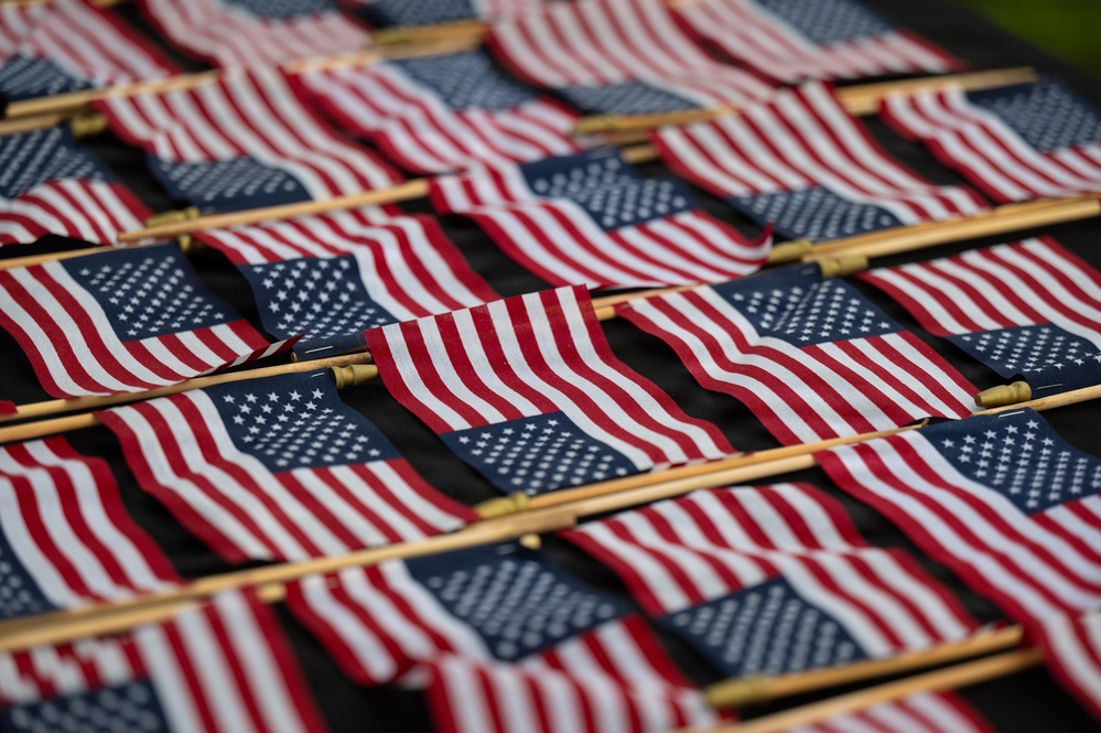 The image shows a large collection of small American flags laid out in rows on a dark surface, with wooden dowel sticks attached. The flags display the characteristic red, white, and blue design with stars and stripes. This appears to be a preparation or staging scene, likely for a patriotic memorial or commemorative event.
