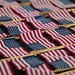 The image shows a large collection of small American flags laid out in rows on a dark surface, with wooden dowel sticks attached. The flags display the characteristic red, white, and blue design with stars and stripes. This appears to be a preparation or staging scene, likely for a patriotic memorial or commemorative event.