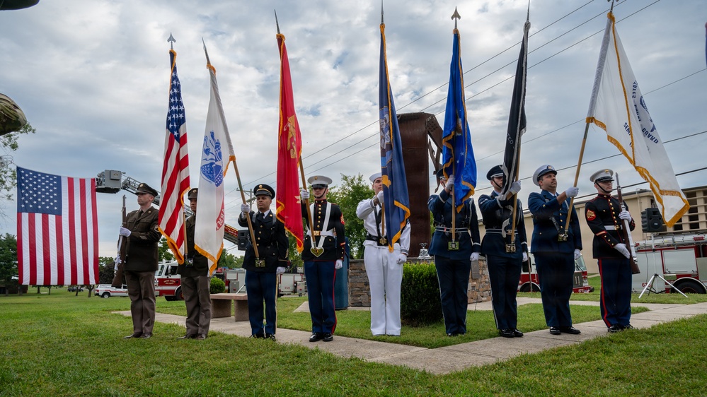 A joint military color guard ceremony is taking place outdoors, with representatives from multiple U.S. armed service branches standing in formation holding their respective service flags and the American flag. The ceremony appears to be held at a memorial or civic venue, with a large American flag displayed on a fire truck ladder in the background. Fire department vehicles and a stone monument are also visible, suggesting a patriotic or memorial observance.