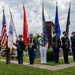 A joint military color guard ceremony is taking place outdoors, with representatives from multiple U.S. armed service branches standing in formation holding their respective service flags and the American flag. The ceremony appears to be held at a memorial or civic venue, with a large American flag displayed on a fire truck ladder in the background. Fire department vehicles and a stone monument are also visible, suggesting a patriotic or memorial observance.