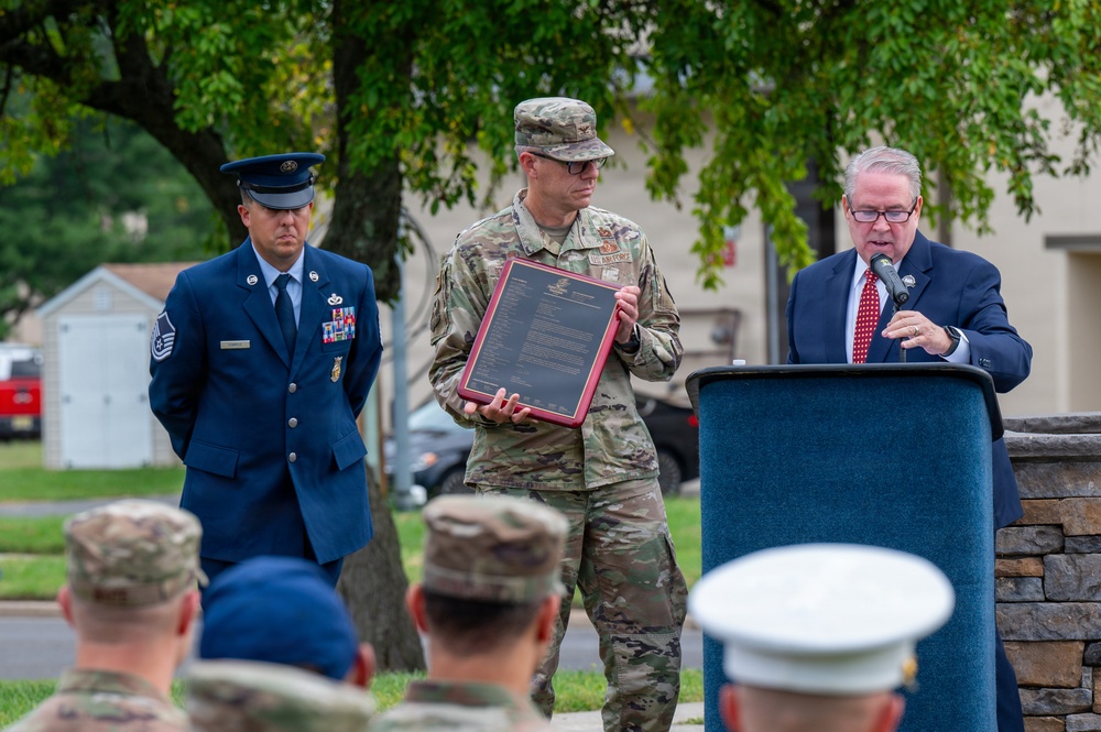 An outdoor military ceremony is taking place, featuring a civilian official speaking at a podium while a soldier in Army Combat Uniform holds a framed plaque or certificate. A senior Air Force enlisted member in dress blues stands nearby, and an audience of mixed military personnel in various uniforms observes from the foreground. The setting appears to be on a military installation grounds.