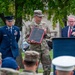 An outdoor military ceremony is taking place, featuring a civilian official speaking at a podium while a soldier in Army Combat Uniform holds a framed plaque or certificate. A senior Air Force enlisted member in dress blues stands nearby, and an audience of mixed military personnel in various uniforms observes from the foreground. The setting appears to be on a military installation grounds.