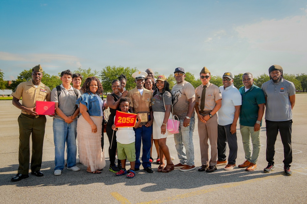 A group of approximately 14 people pose together on what appears to be a military parade ground or tarmac during a Marine Corps graduation or family day event. A central figure in Marine dress uniform with a white cover (hat) is surrounded by family members and friends, with one person holding a red pennant marked '2056' and another holding a red box and a plaque. The occasion appears to be a celebratory military milestone, likely a boot camp graduation.