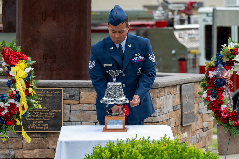 A U.S. Air Force Staff Sergeant in dress blues is ringing a ceremonial silver bell at what appears to be a 9/11 memorial ceremony. The memorial features a stone wall with a '9/11' plaque, a rusted steel beam (likely from the World Trade Center), and patriotic floral wreaths. The setting suggests a formal commemorative event at Joint Base McGuire-Dix-Lakehurst (JB MDL), as indicated by text visible on the donor plaque.