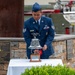 A U.S. Air Force Staff Sergeant in dress blues is ringing a ceremonial silver bell at what appears to be a 9/11 memorial ceremony. The memorial features a stone wall with a '9/11' plaque, a rusted steel beam (likely from the World Trade Center), and patriotic floral wreaths. The setting suggests a formal commemorative event at Joint Base McGuire-Dix-Lakehurst (JB MDL), as indicated by text visible on the donor plaque.