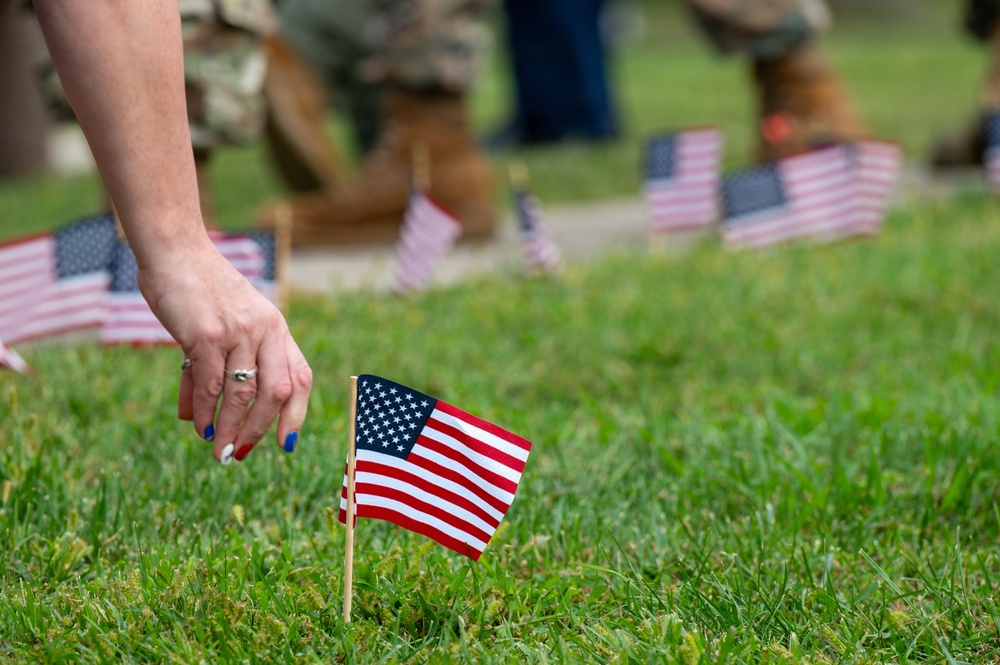 A person is placing or adjusting a small American flag in a grass lawn, with multiple other small American flags already planted in rows across the field. In the blurred background, individuals wearing military camouflage uniforms are visible, also placing flags. This appears to be a memorial or patriotic ceremony, likely associated with Veterans Day, Memorial Day, or a similar commemorative event.
