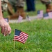A person is placing or adjusting a small American flag in a grass lawn, with multiple other small American flags already planted in rows across the field. In the blurred background, individuals wearing military camouflage uniforms are visible, also placing flags. This appears to be a memorial or patriotic ceremony, likely associated with Veterans Day, Memorial Day, or a similar commemorative event.