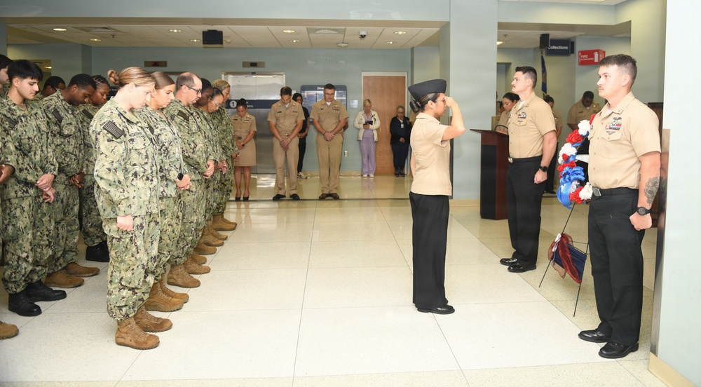 A military ceremony is taking place inside what appears to be a government or military building, likely a memorial or commemorative event. Personnel in Navy working uniforms (NWUs) and khaki service uniforms are assembled in formation, with one service member rendering a salute. A red, white, and blue floral wreath is prominently displayed, suggesting a patriotic or memorial observance.