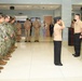 A military ceremony is taking place inside what appears to be a government or military building, likely a memorial or commemorative event. Personnel in Navy working uniforms (NWUs) and khaki service uniforms are assembled in formation, with one service member rendering a salute. A red, white, and blue floral wreath is prominently displayed, suggesting a patriotic or memorial observance.