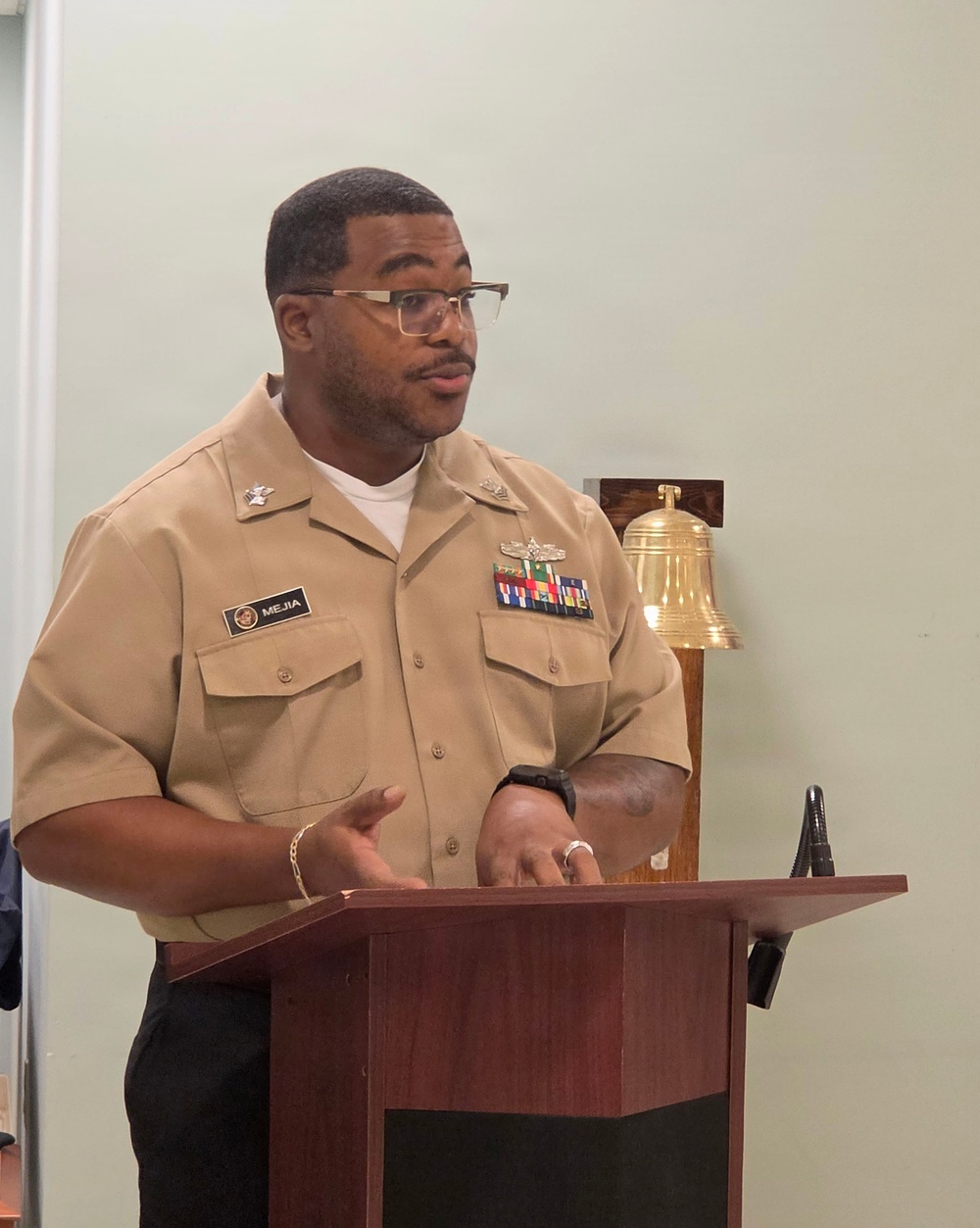 A U.S. Navy enlisted service member named Mejia is speaking at a wooden podium, wearing the tan Service Uniform (Type III). The individual displays multiple service ribbons, a warfare device badge, and rank insignia on their uniform. A brass ship's bell on a wooden mount is visible in the background.
