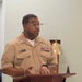 A U.S. Navy enlisted service member named Mejia is speaking at a wooden podium, wearing the tan Service Uniform (Type III). The individual displays multiple service ribbons, a warfare device badge, and rank insignia on their uniform. A brass ship's bell on a wooden mount is visible in the background.