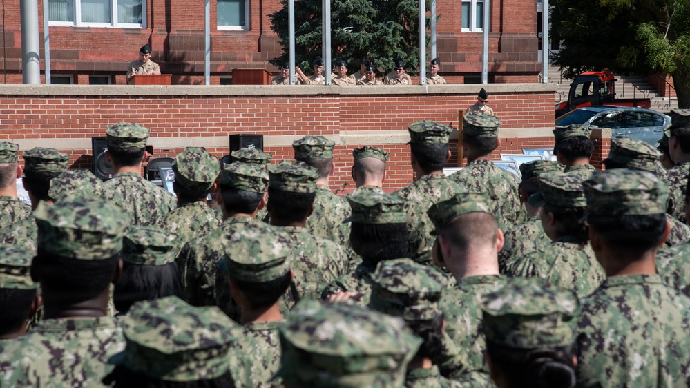 A large formation of military personnel in camouflage uniforms are assembled outdoors facing a raised platform or elevated area where a speaker stands at a podium. The setting appears to be a military base courtyard with a red brick building in the background. This appears to be a formal military ceremony, briefing, or address event.
