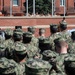 A large formation of military personnel in camouflage uniforms are assembled outdoors facing a raised platform or elevated area where a speaker stands at a podium. The setting appears to be a military base courtyard with a red brick building in the background. This appears to be a formal military ceremony, briefing, or address event.