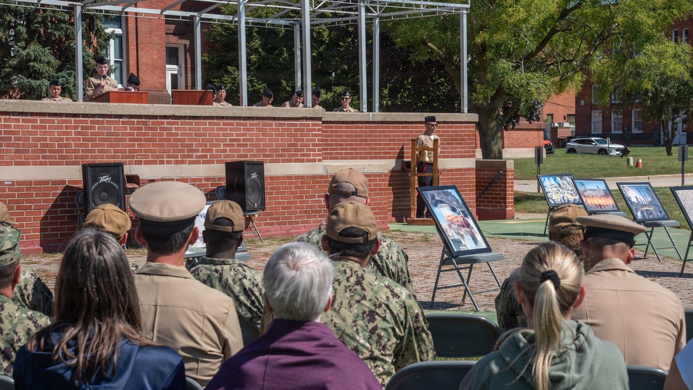 An outdoor military ceremony is taking place at what appears to be a military installation, with uniformed personnel and civilians seated as an audience facing a speaker at a podium. Several framed photographs are displayed on easels to the right, and a raised platform with additional personnel is visible in the background. The brick architecture and formal arrangement suggest a commemorative or memorial event.