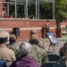 An outdoor military ceremony is taking place at what appears to be a military installation, with uniformed personnel and civilians seated as an audience facing a speaker at a podium. Several framed photographs are displayed on easels to the right, and a raised platform with additional personnel is visible in the background. The brick architecture and formal arrangement suggest a commemorative or memorial event.