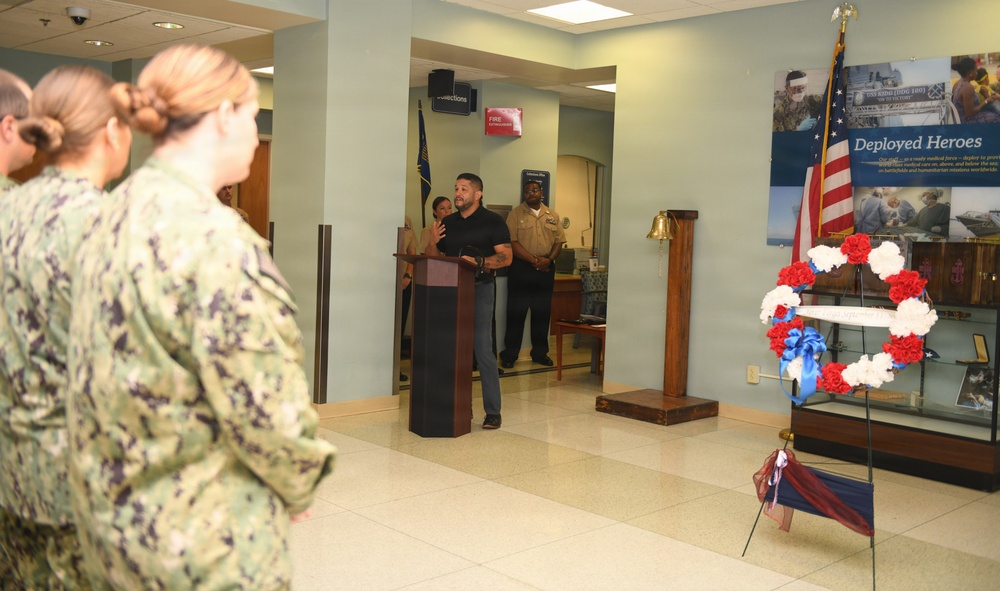 A military ceremony is taking place in what appears to be a naval facility lobby or common area. A civilian man in a black shirt is speaking at a podium while uniformed Navy personnel stand at attention. The setting includes patriotic decorations including a red, white, and blue floral wreath and an American flag, alongside a 'Deployed Heroes' display board.