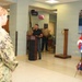 A military ceremony is taking place in what appears to be a naval facility lobby or common area. A civilian man in a black shirt is speaking at a podium while uniformed Navy personnel stand at attention. The setting includes patriotic decorations including a red, white, and blue floral wreath and an American flag, alongside a 'Deployed Heroes' display board.