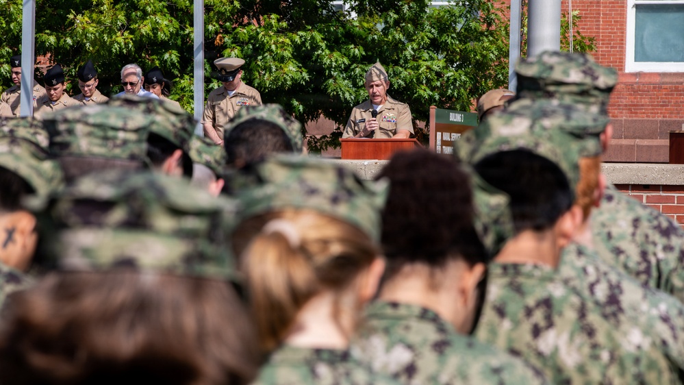 A U.S. Navy officer in Service Khaki uniform is speaking at a podium during an outdoor military ceremony. The audience consists of personnel wearing Navy Working Uniform (NWU) camouflage pattern, viewed from behind in the foreground. A sign partially visible in the background references 'Naval Service' and 'Building 3', suggesting a training command facility.