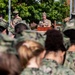 A U.S. Navy officer in Service Khaki uniform is speaking at a podium during an outdoor military ceremony. The audience consists of personnel wearing Navy Working Uniform (NWU) camouflage pattern, viewed from behind in the foreground. A sign partially visible in the background references 'Naval Service' and 'Building 3', suggesting a training command facility.