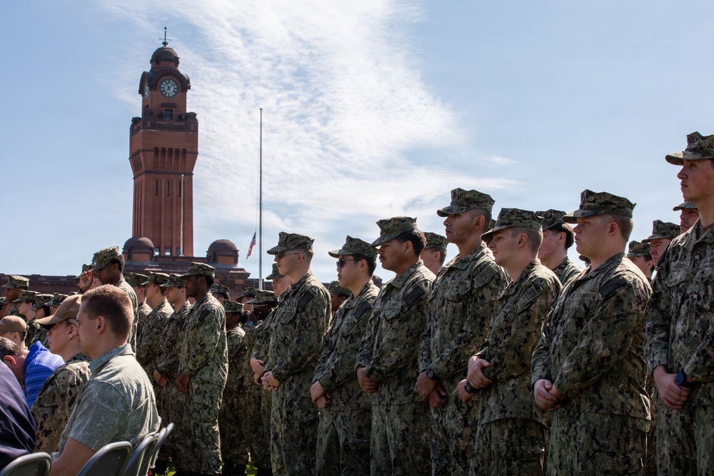 A large formation of U.S. Navy personnel in NWU Type III (woodland digital camouflage) uniforms stand at attention during what appears to be an outdoor military ceremony or formation. In the background, a prominent red-brick clock tower and a flagpole with a flag at half-staff are visible. Civilian observers are seated to the left, suggesting this is a formal commemorative or official event.